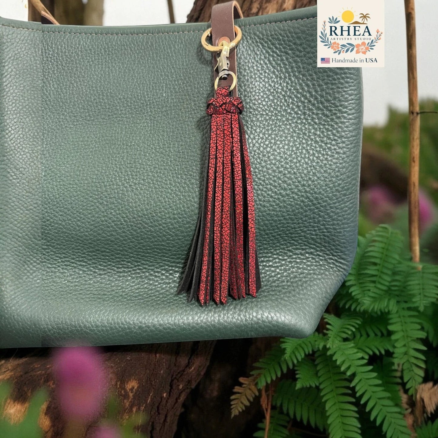Rhea Artistry Studio handmade dual-tone leather tassel keychain in red and black with brushed brass clasp attached to a green pebbled leather bag.