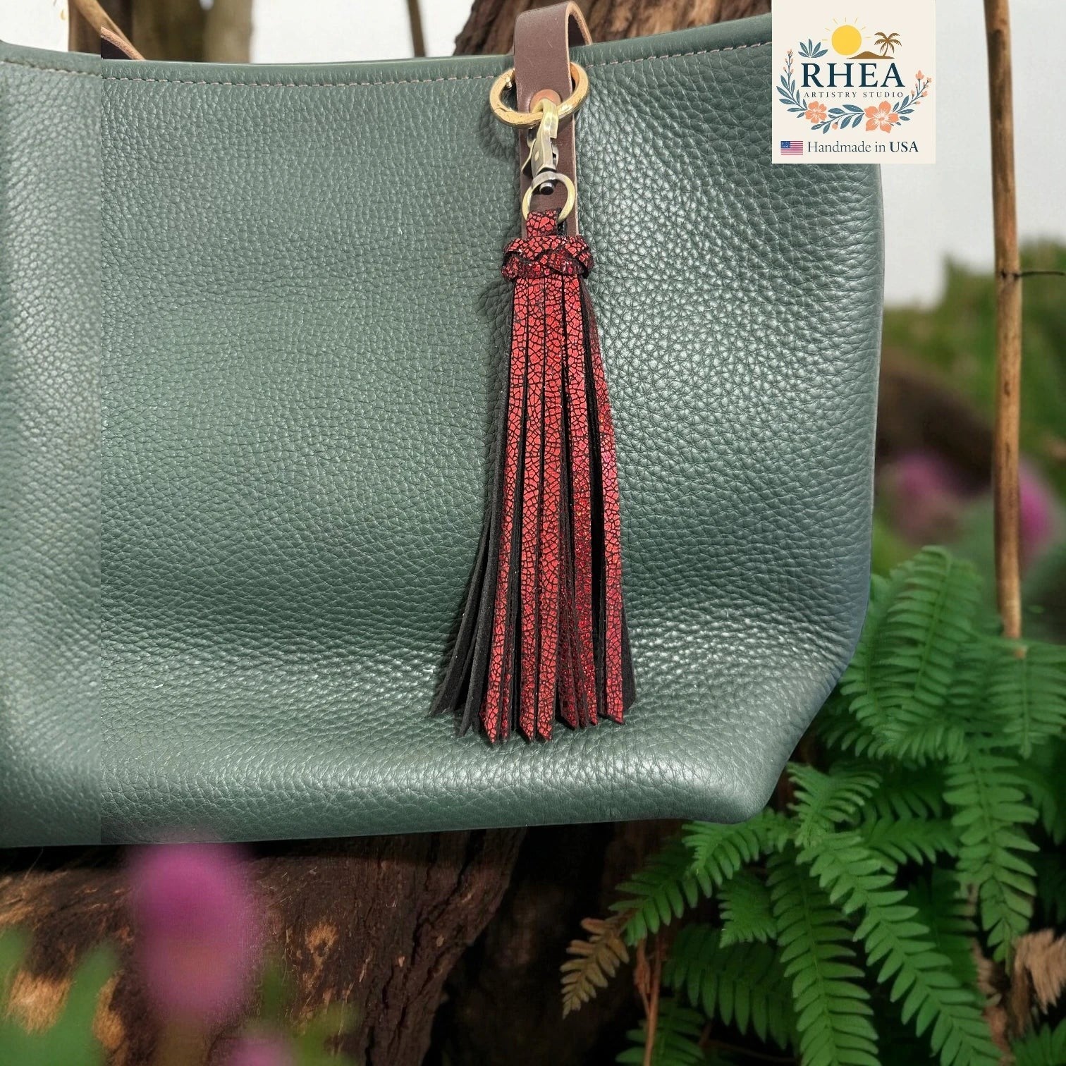 Rhea Artistry Studio handmade dual-tone leather tassel keychain in red and black with brushed brass clasp attached to a green pebbled leather bag.