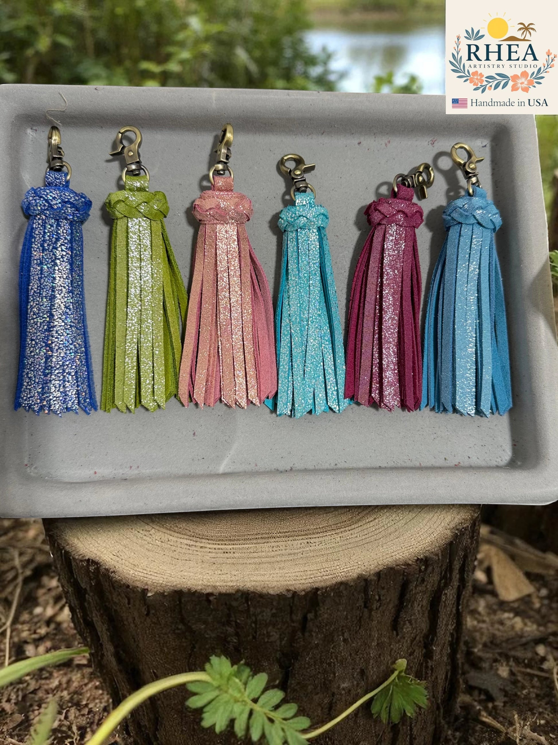 Rhea Artistry Studio handmade leather tassel keychains in blue, lime green, pink, teal, magenta, and light blue with glitter accents and lobster clasps.
