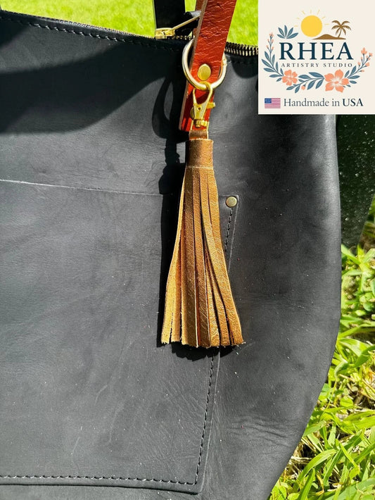 Rhea Artistry Studio tan full-grain leather jumbo tassel keychain with lobster clasp attached to a black leather tote bag.