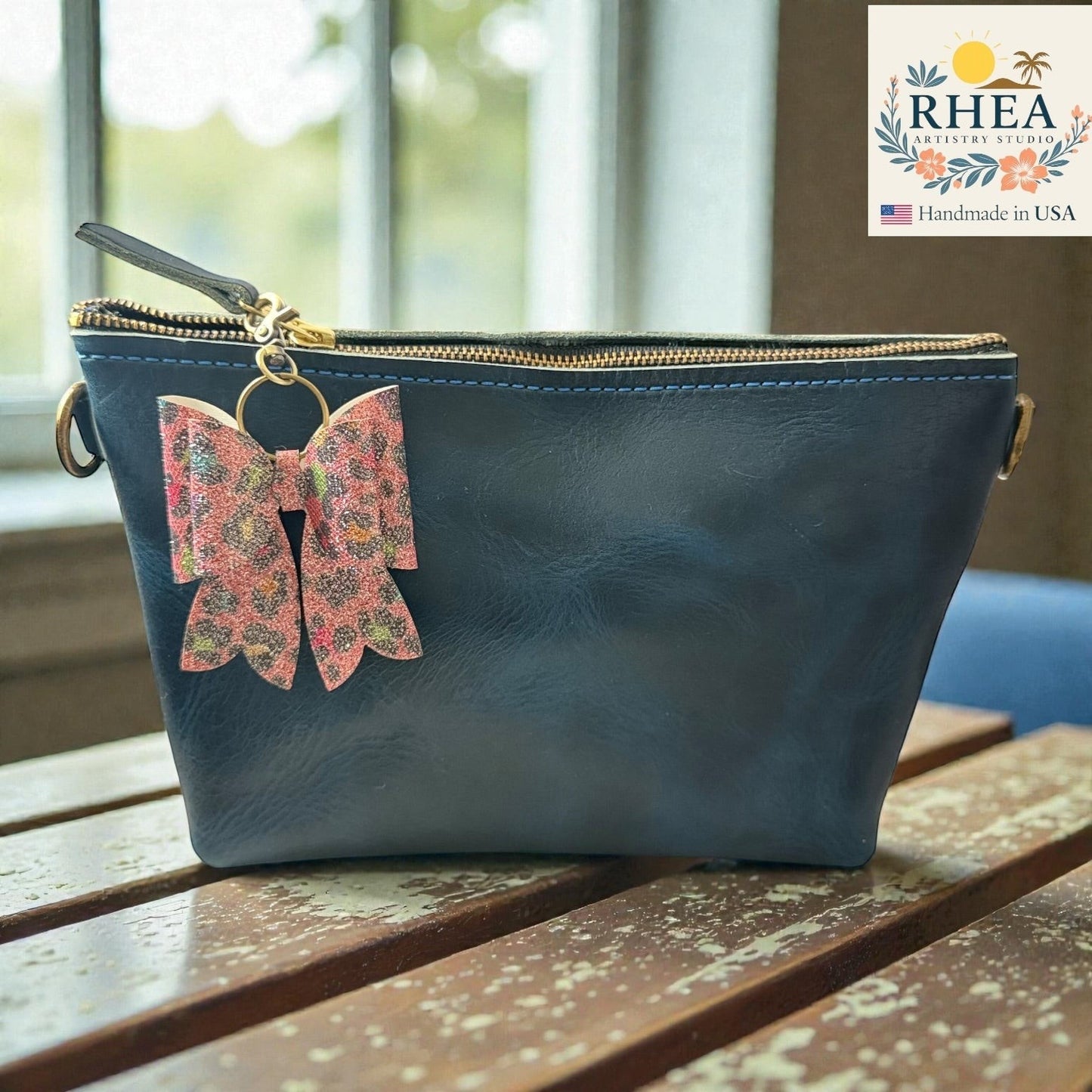 Rhea Artistry Studio handmade pink leopard print bow bag charm with brass hardware attached to a navy blue leather pouch.