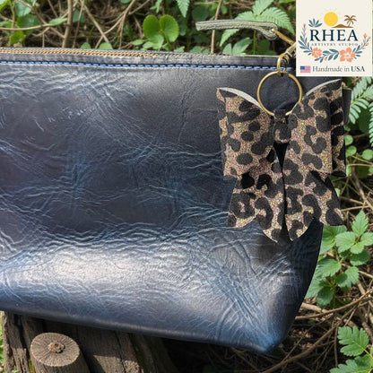 Rhea Artistry Studio handmade leopard print bow bag charm with brass hardware, clipped to a dark blue leather pouch.