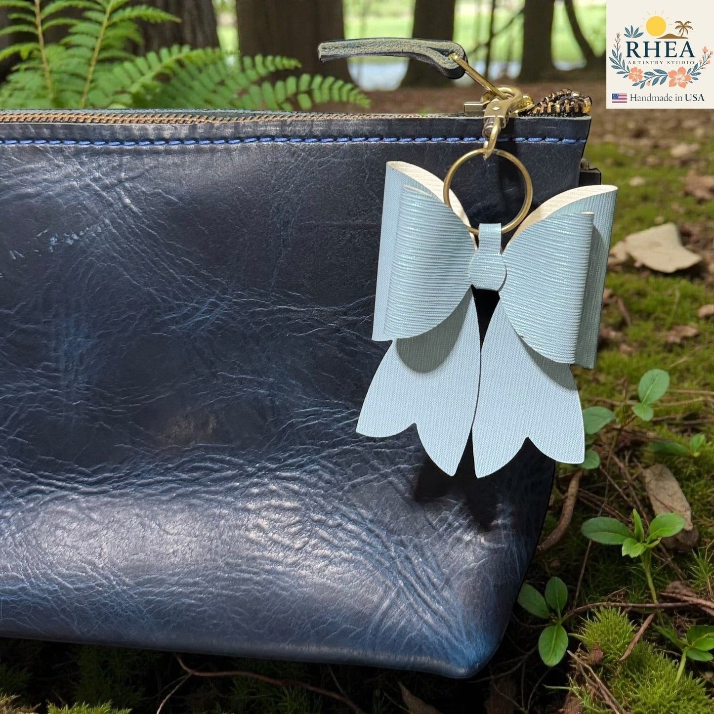 Rhea Artistry Studio light blue double-layer French bow bag charm with brass hardware attached to a dark blue textured leather pouch.
