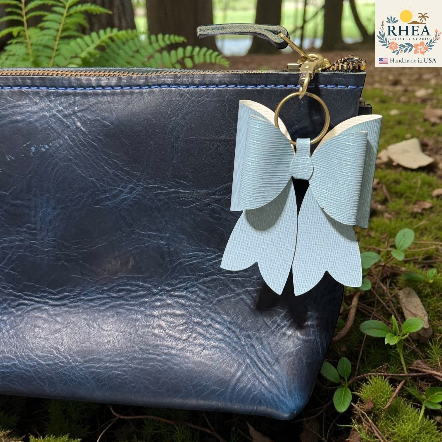 Rhea Artistry Studio light blue double-layer French bow bag charm with brass hardware attached to a dark blue textured leather pouch.