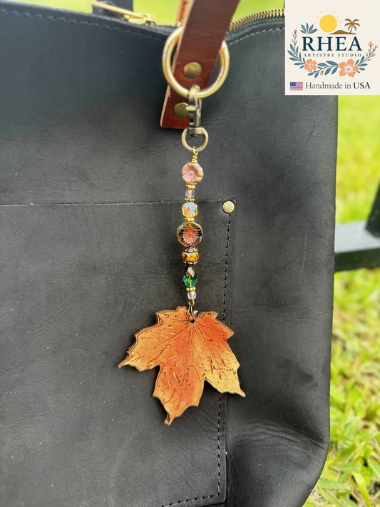 Rhea Artistry Studio orange maple leaf charm with colorful beads and brass hardware, attached to a black leather bag.