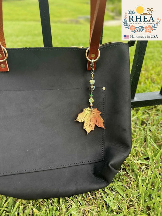 Rhea Artistry Studio handcrafted maple leaf charm with green beads on a black leather tote bag.