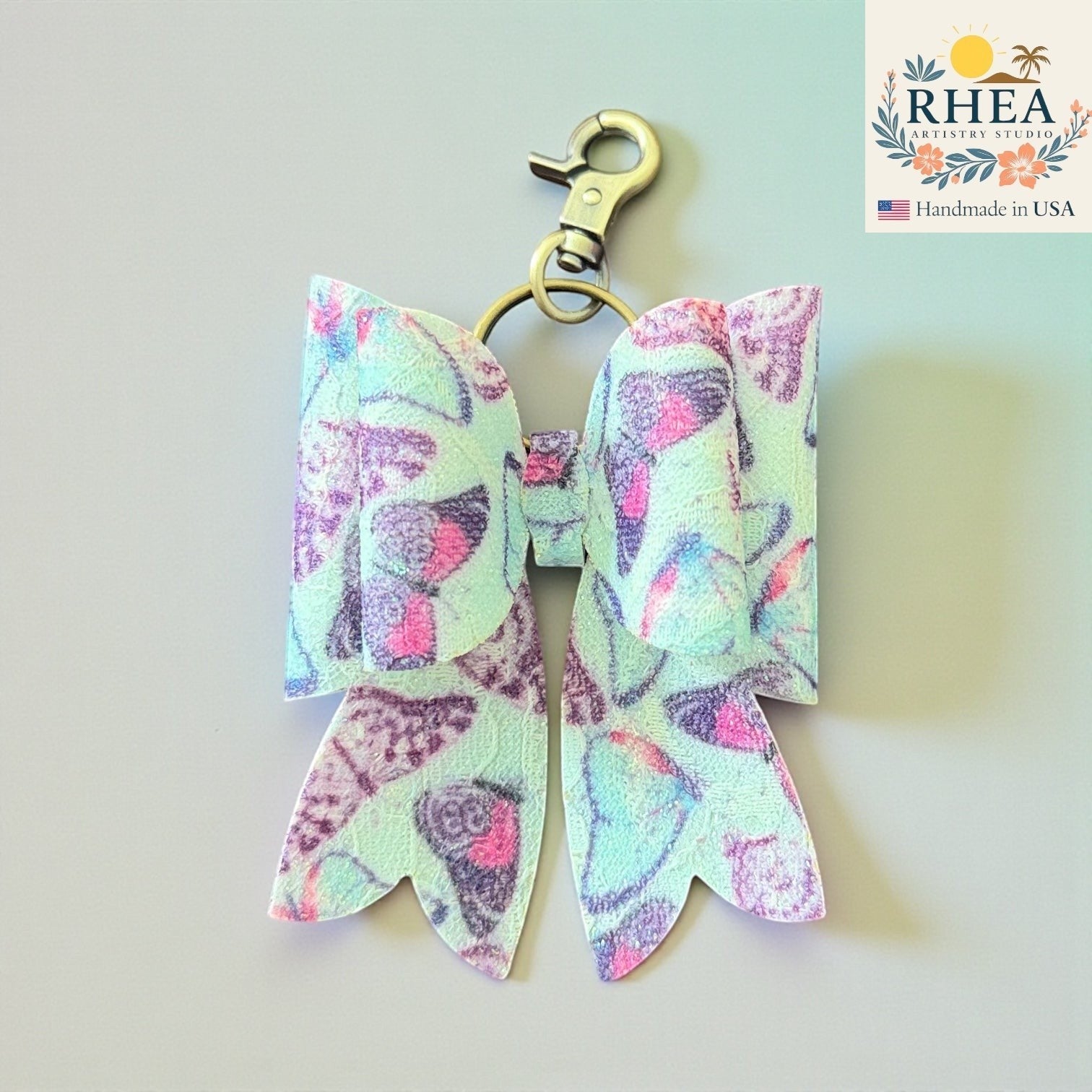 Rhea Artistry Studio handmade mint green bow charm with purple and pink butterfly print and brass clasp.