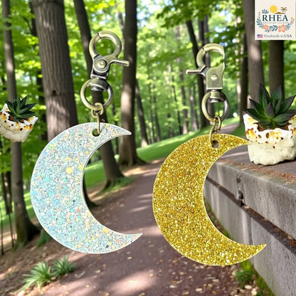 Rhea Artistry Studio gold glitter and holographic glitter moon acrylic charms with brass clasps, displayed outdoors.