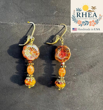 Rhea Artistry Studio mustard hibiscus drop earrings with brass accents and orange beads, approx. 1.5-2 inches long.