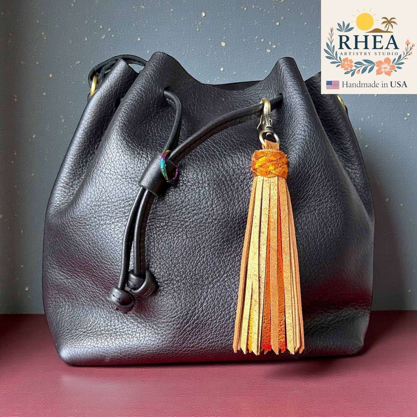Rhea Artistry Studio handmade orange metallic leather tassel charm attached to a black leather drawstring bag.