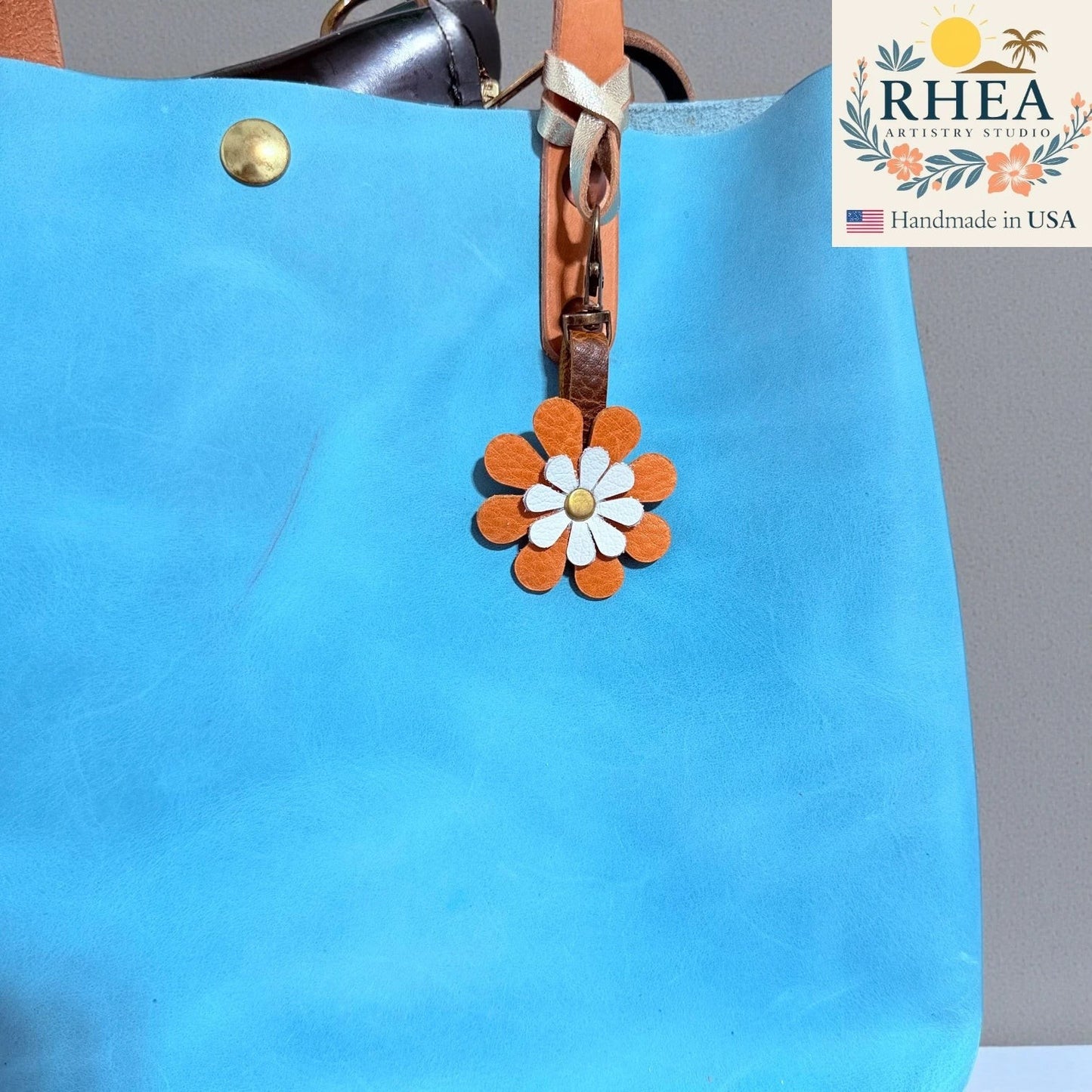 Rhea Artistry Studio handmade double-layer leather daisy charm in orange and white with a brass clasp, attached to a blue tote bag.