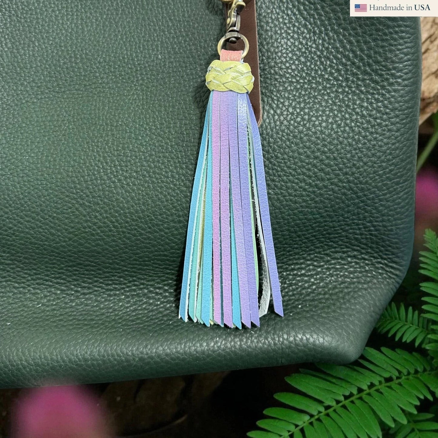 Rhea Artistry Studio pastel leather tassel keychain with braided yellow top and lobster clasp attached to a dark green pebbled leather bag.