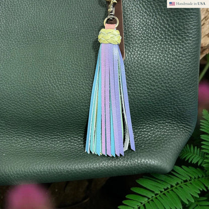 Rhea Artistry Studio pastel leather tassel keychain with braided yellow top and lobster clasp attached to a dark green pebbled leather bag.