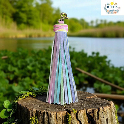 Rhea Artistry Studio pastel rainbow leather tassel keychain with lobster clasp, displayed on a tree stump outdoors.