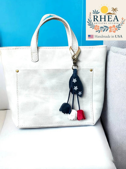 Rhea Artistry Studio patriotic leather flower charm with red, white, and blue tulips and silver stars, attached to a white tote bag.