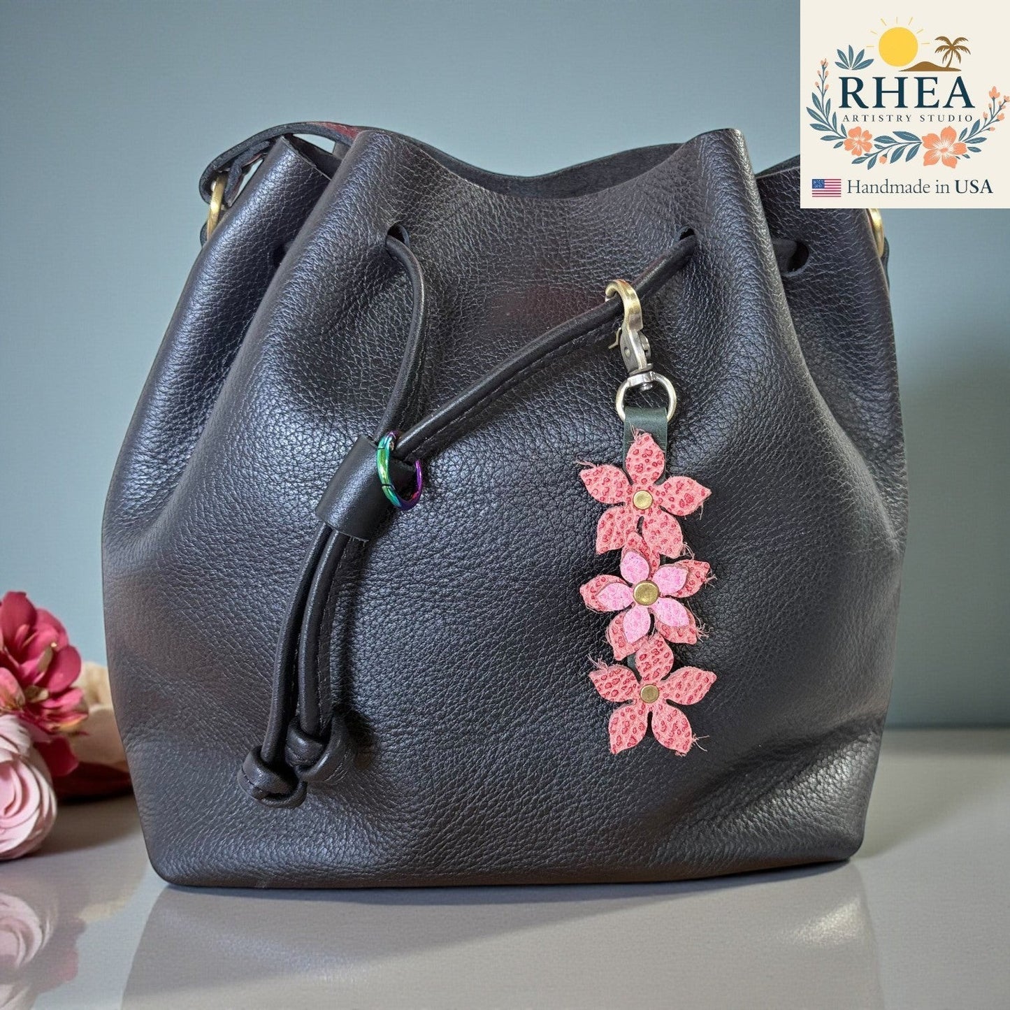 Rhea Artistry Studio handmade pink floral leather charm with brushed brass clasp attached to a black leather drawstring bag.