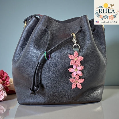 Rhea Artistry Studio handmade pink floral leather charm with brushed brass clasp attached to a black leather drawstring bag.