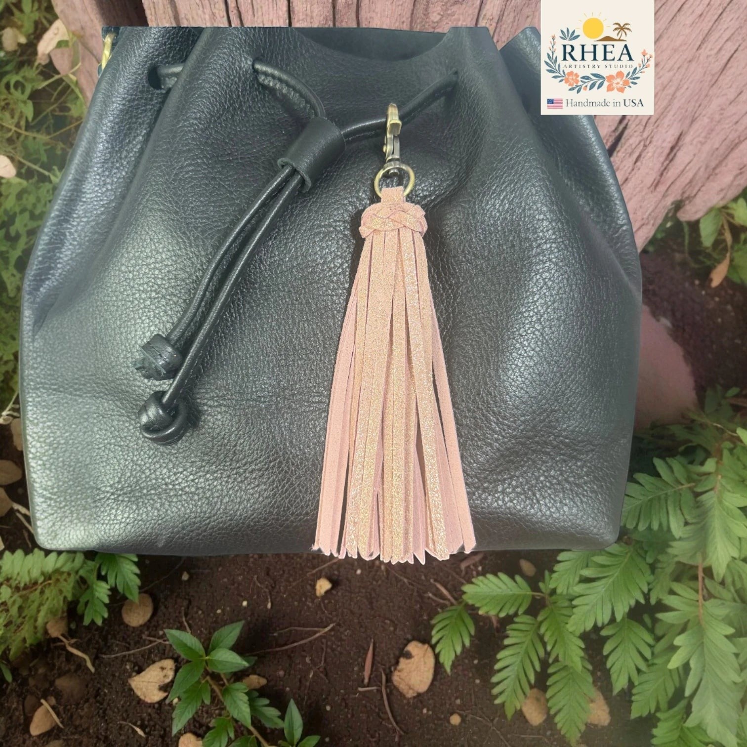 Rhea Artistry Studio handmade light pink glitter leather tassel keychain with lobster clasp attached to a black leather drawstring bag.