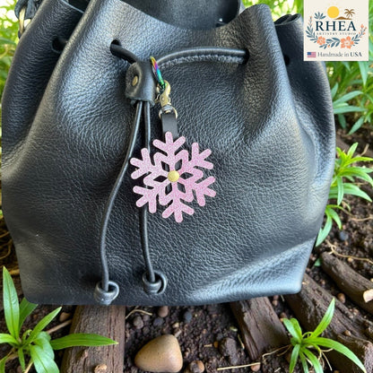 Rhea Artistry Studio pink glitter snowflake leather charm with gold center, clipped to a black leather drawstring bag.