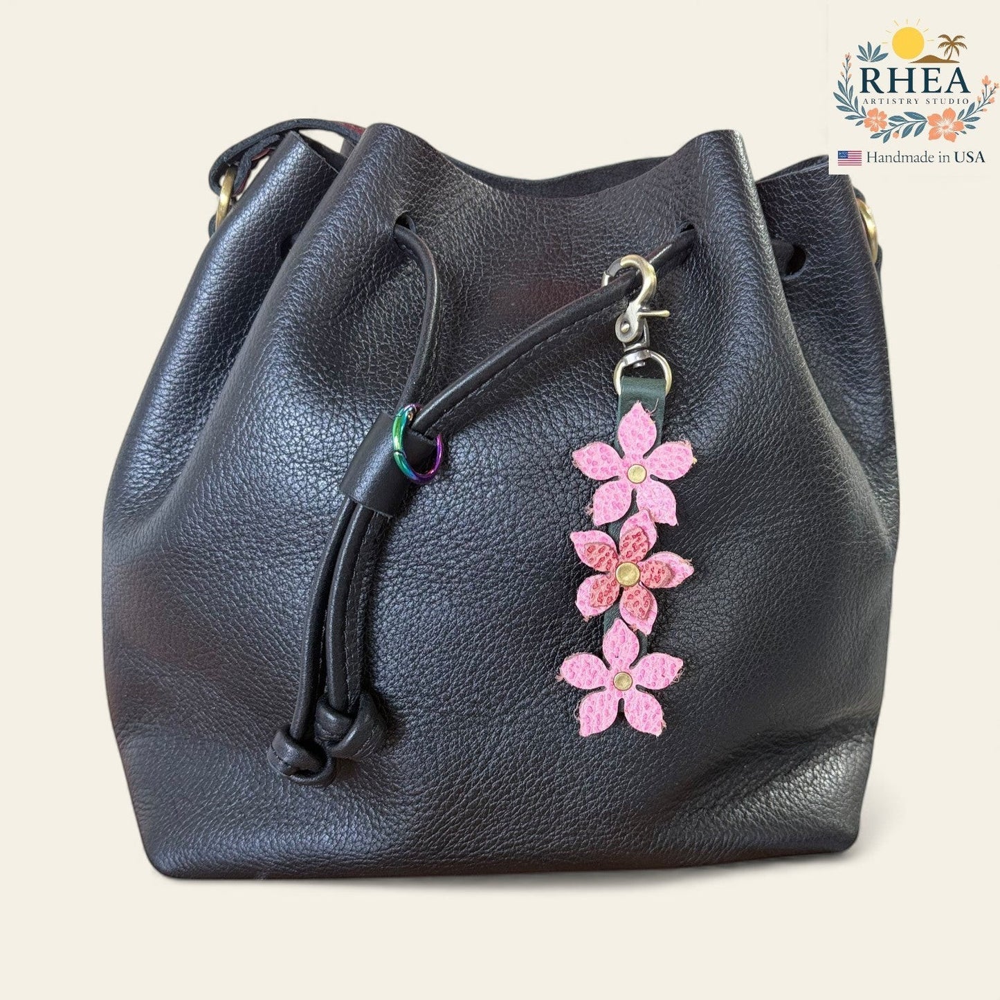 Rhea Artistry Studio handmade pink leather floral charm with brushed brass clasp attached to a black leather bucket bag.