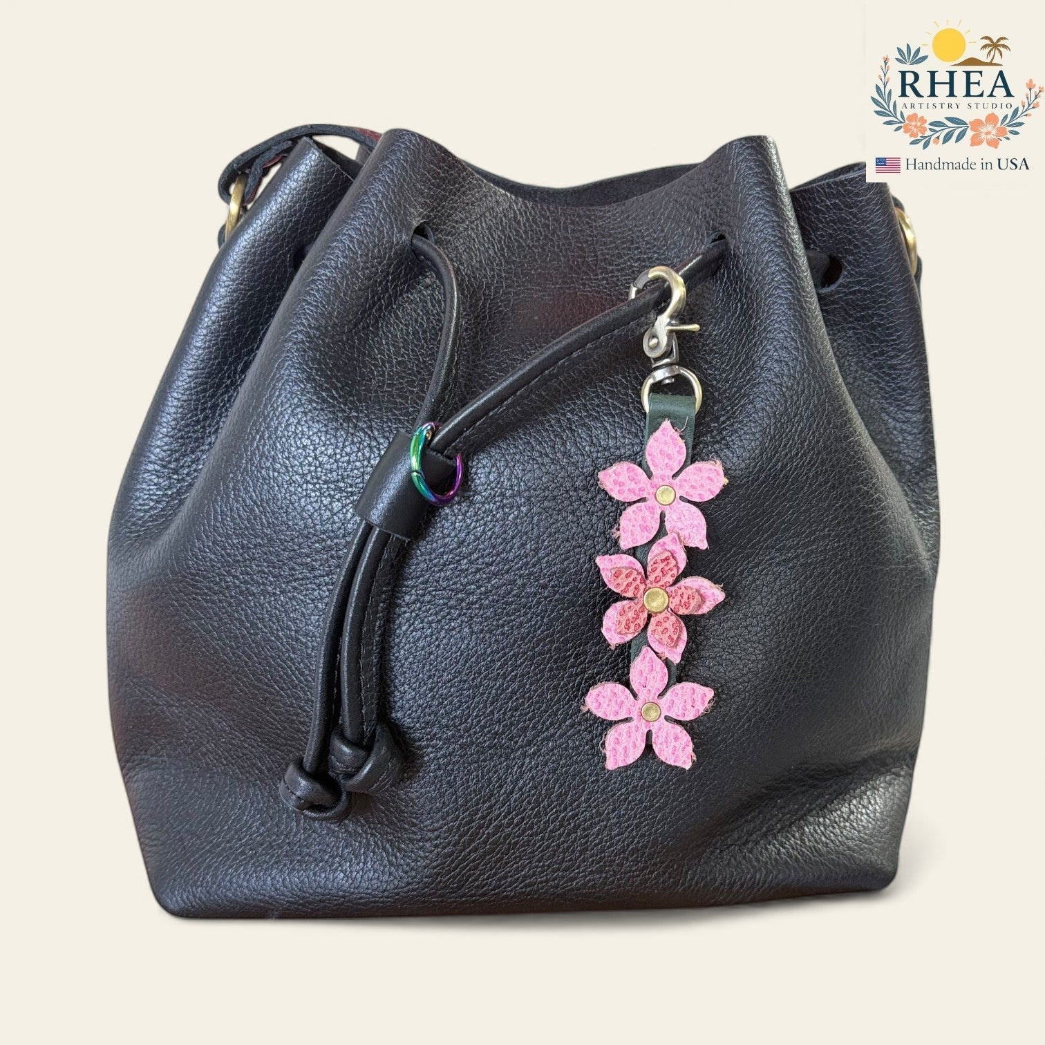 Rhea Artistry Studio handmade pink leather floral charm with brushed brass clasp attached to a black leather bucket bag.