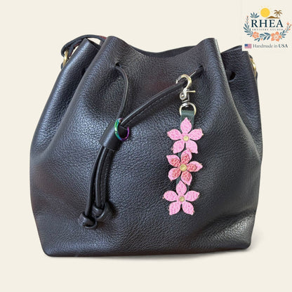 Rhea Artistry Studio handmade pink leather floral charm with brushed brass clasp attached to a black leather bucket bag.