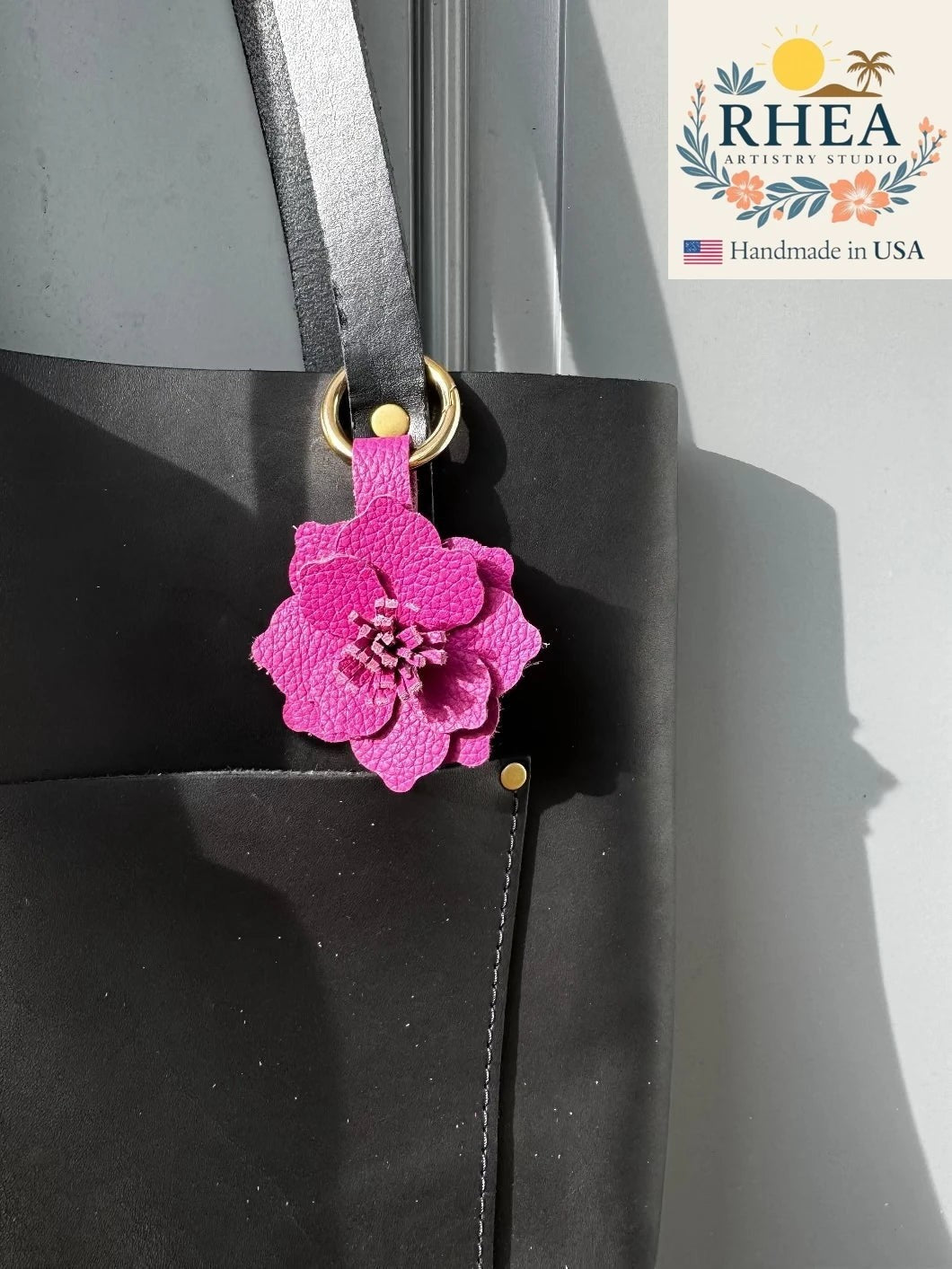Rhea Artistry Studio pink leather hibiscus flower charm with split ring and lobster clasp attached to a black tote bag.