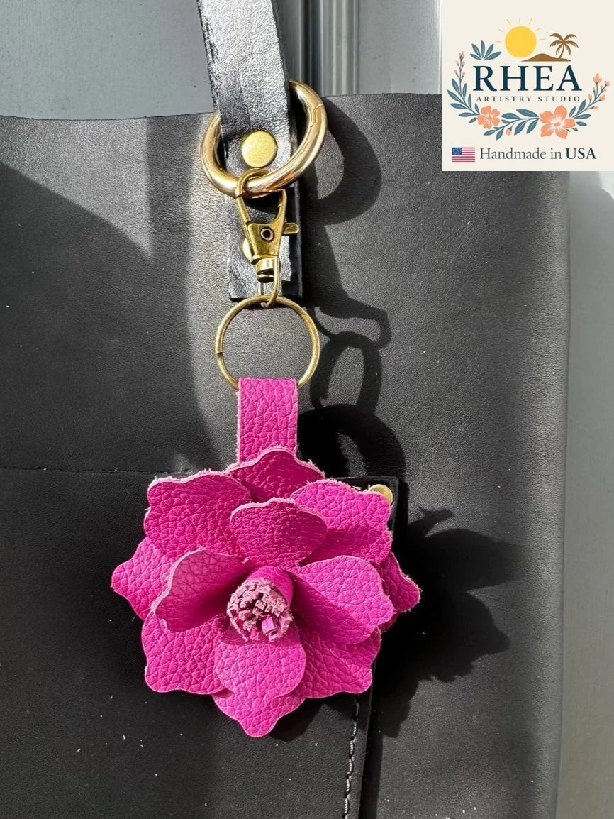 Rhea Artistry Studio pink hibiscus leather flower charm with gold clasp and keyring attached to a black tote bag.