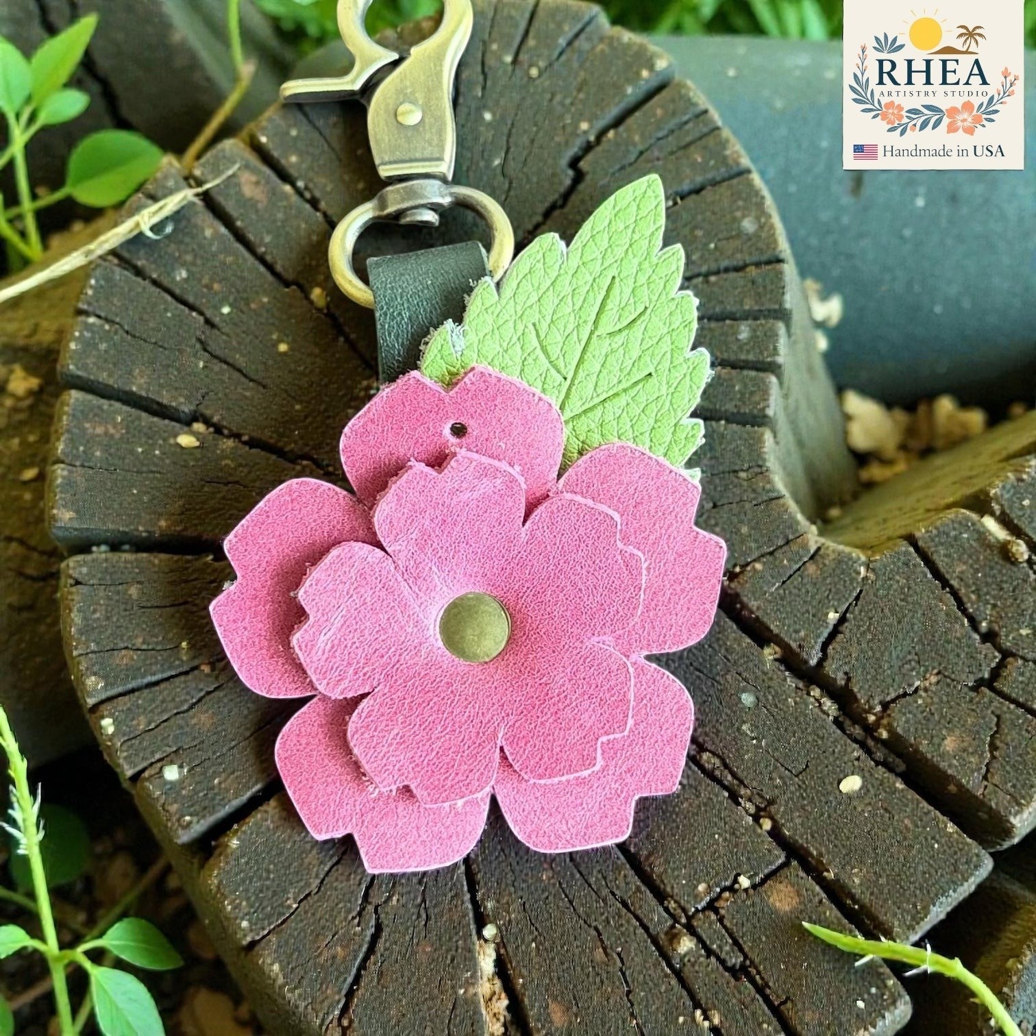 Rhea Artistry Studio pink layered leather rose charm with green engraved leaf and brushed brass hardware, attached to a dark wood surface.