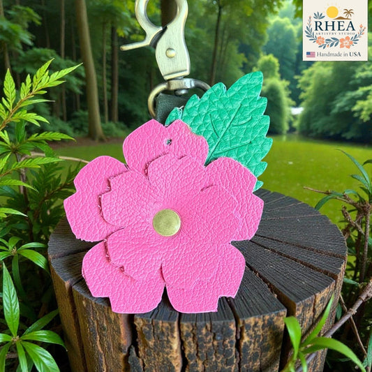 Rhea Artistry Studio pink layered leather rose charm with green engraved leaf and brass hardware, attached to a dark wood stump.