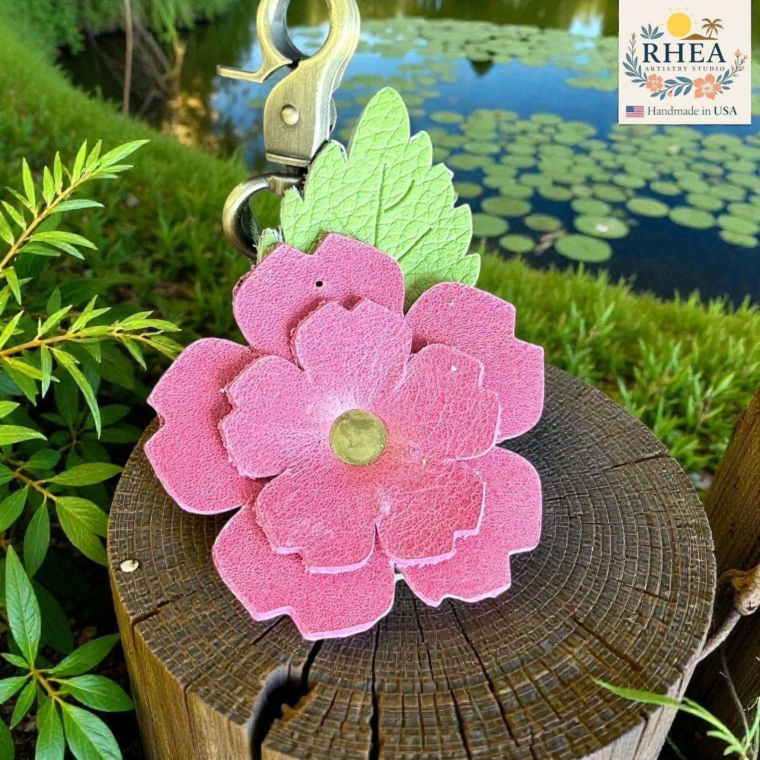 Rhea Artistry Studio pink layered leather rose charm with engraved green leaf and brass hardware, attached to a wooden log.