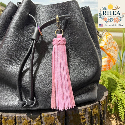 Rhea Artistry Studio pink leather tassel bag charm with braided detail and brass clasp, attached to a black leather bag.