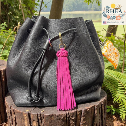 Rhea Artistry Studio bright pink leather tassel bag charm with braided detail and brass clasp on a black leather bag.