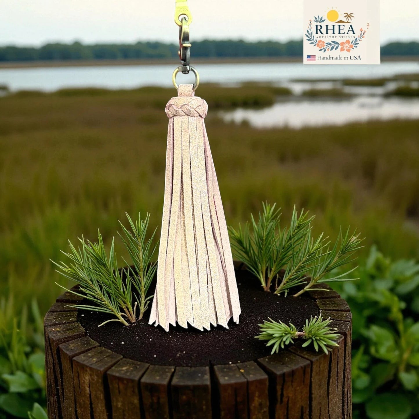 Rhea Artistry Studio handmade light pink leather tassel keychain with braided top and lobster clasp, displayed on a wooden post with greenery.