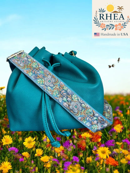 Rhea Artistry Studio 2-inch wide crossbody strap with plum floral embroidery on a teal bucket bag in a field of wildflowers.