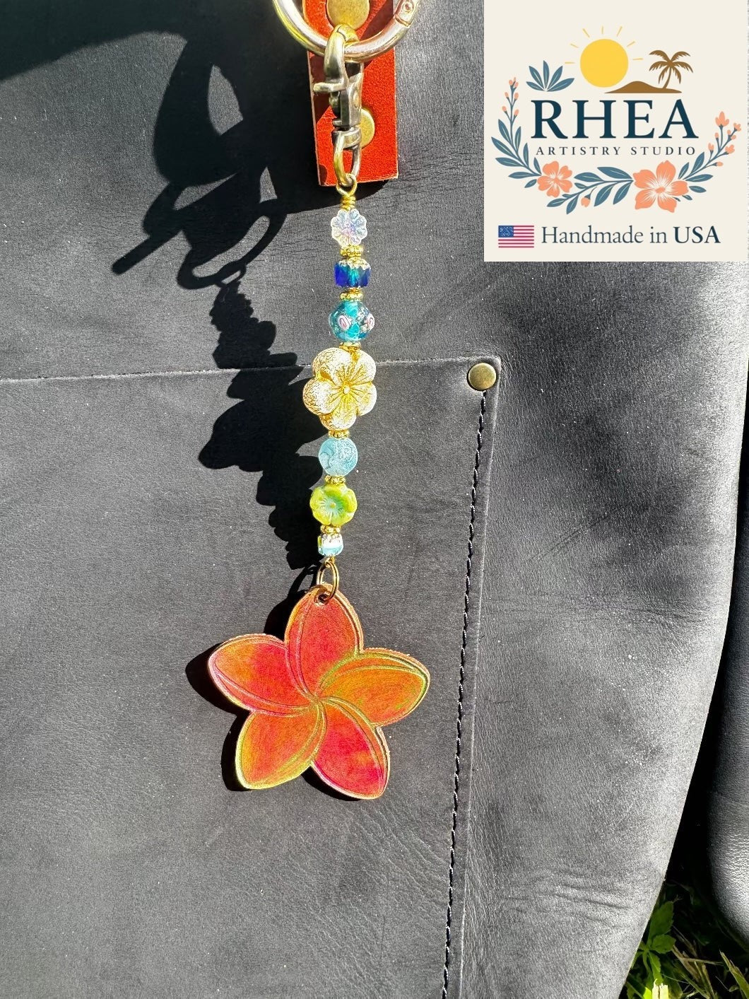 Rhea Artistry Studio Plumeria Whisper charm with a red and orange plumeria pendant and colorful glass beads on a black leather bag.