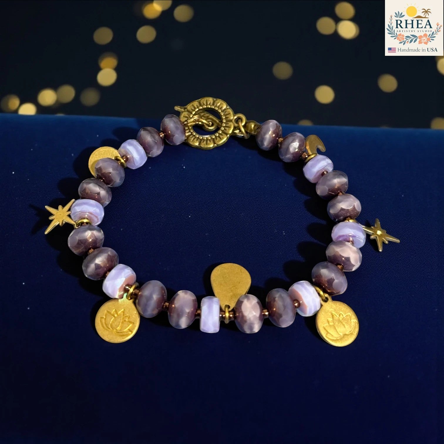 Rhea Artistry Studio handmade bracelet with purple glass beads, brass celestial charms including stars, moons, and lotus motifs, and a floral toggle clasp.