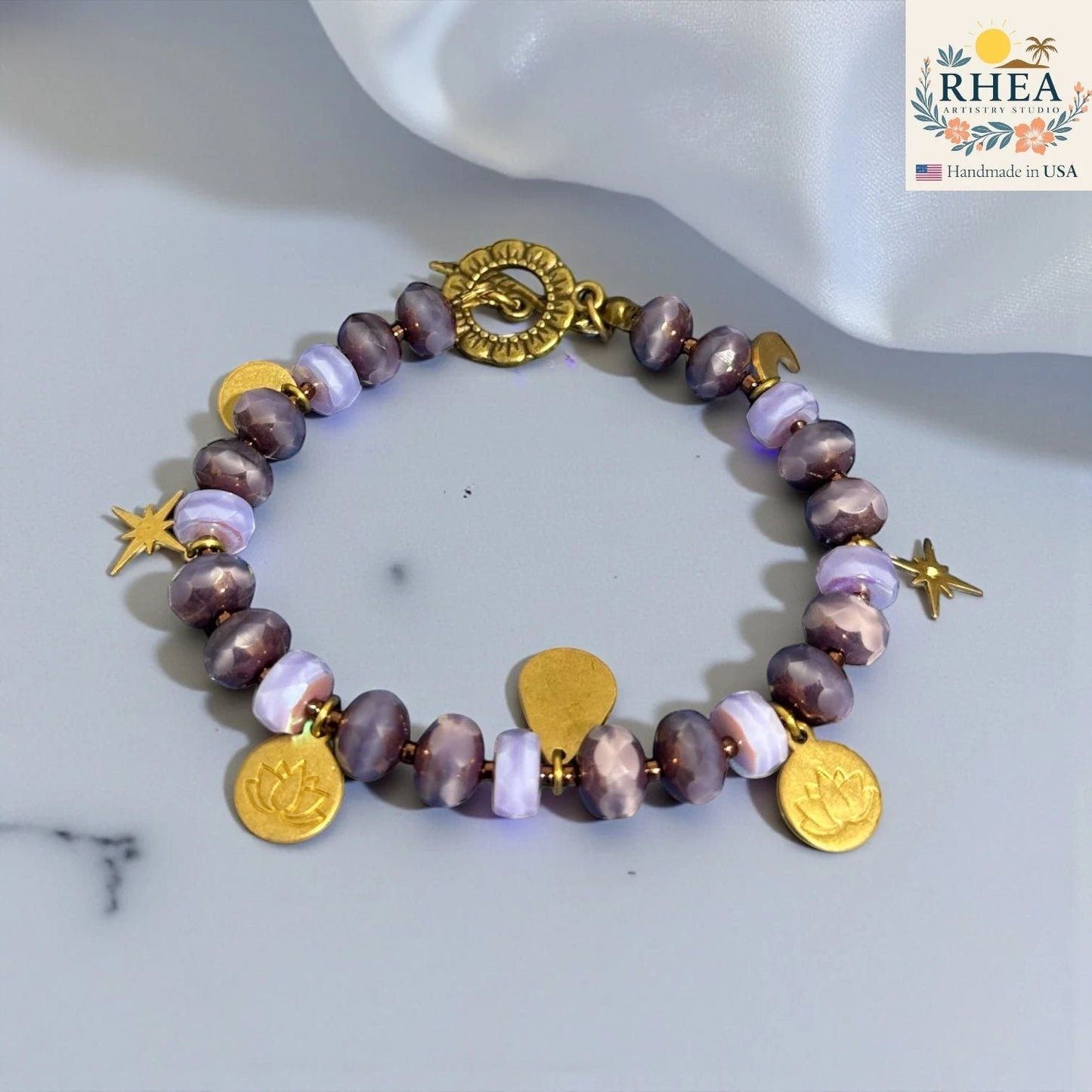 Rhea Artistry Studio handmade bracelet with purple glass beads, brass celestial charms including stars, moons, and lotus motifs, and a floral toggle clasp.
