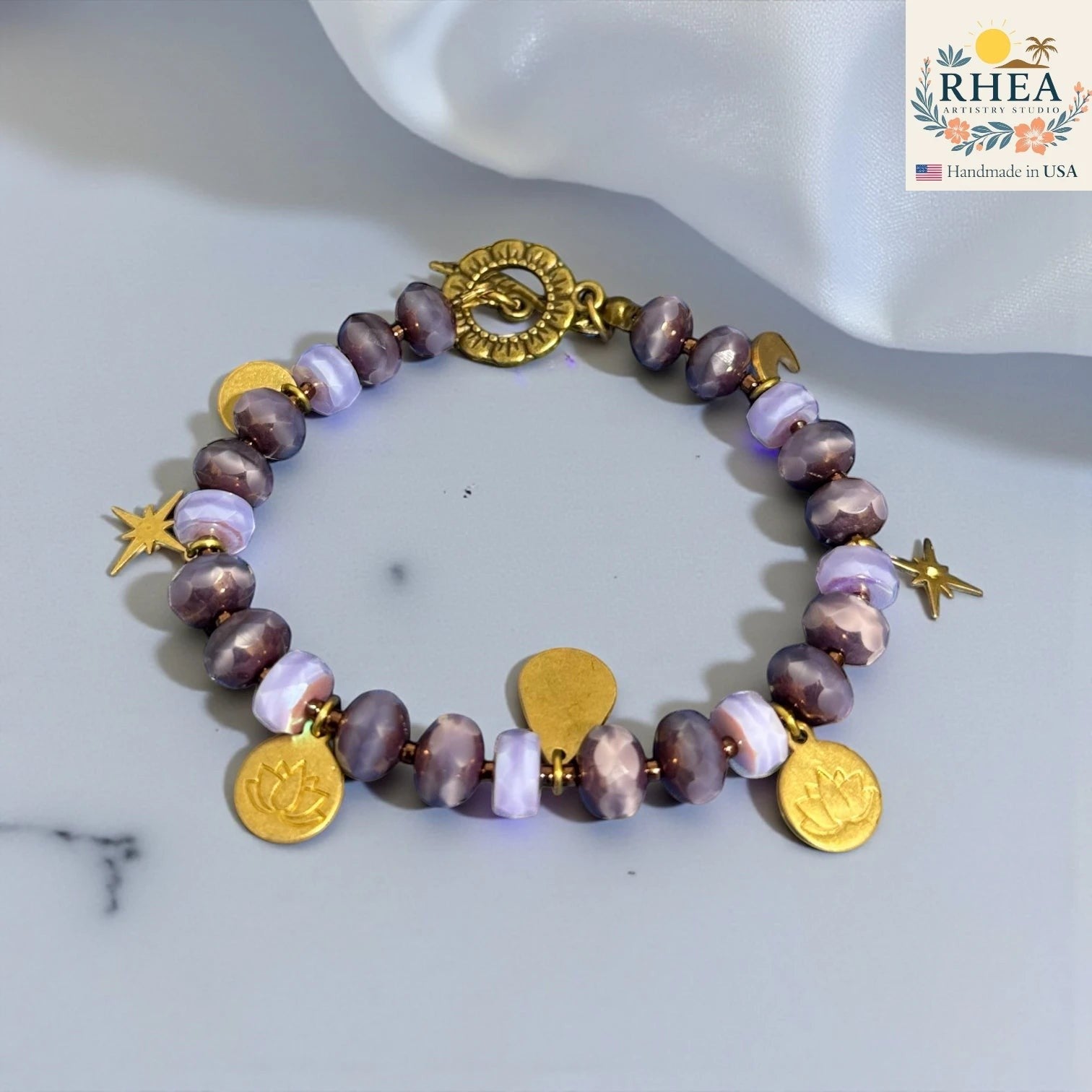 Rhea Artistry Studio handmade bracelet with purple glass beads, brass celestial charms including stars, moons, and lotus motifs, and a floral toggle clasp.