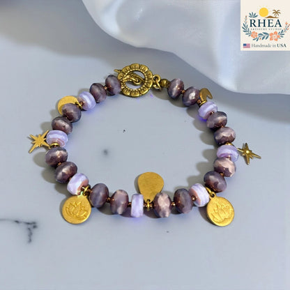Rhea Artistry Studio handmade bracelet with purple glass beads, brass celestial charms including stars, moons, and lotus motifs, and a floral toggle clasp.
