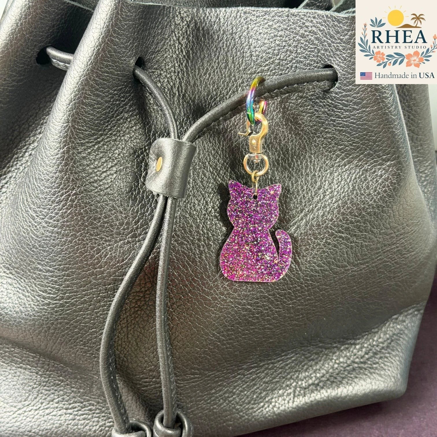 Rhea Artistry Studio purple glitter cat silhouette acrylic charm with antique brass clasp on a black leather bag.