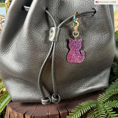Rhea Artistry Studio purple glitter cat silhouette acrylic charm with antique brass clasp on a black leather bag.