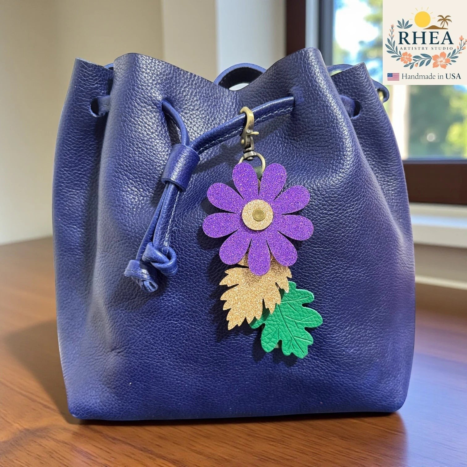 Rhea Artistry Studio purple glitter flower charm with gold and green embossed leather leaves on a blue drawstring bag.