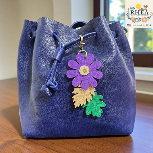 Rhea Artistry Studio purple glitter flower charm with gold and green embossed leather leaves on a blue drawstring bag.