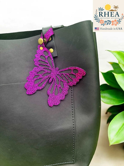 Rhea Artistry Studio purple leather butterfly charm with metallic iridescent finish attached to a black tote bag.