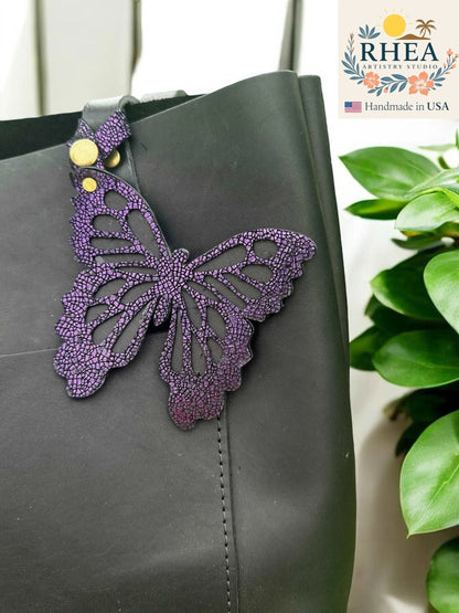 Rhea Artistry Studio purple leather butterfly charm with metallic iridescent finish attached to a black tote bag.