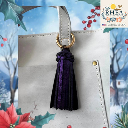 Rhea Artistry Studio handmade dark purple leather tassel bag charm with gold lobster clasp attached to a white tote bag.