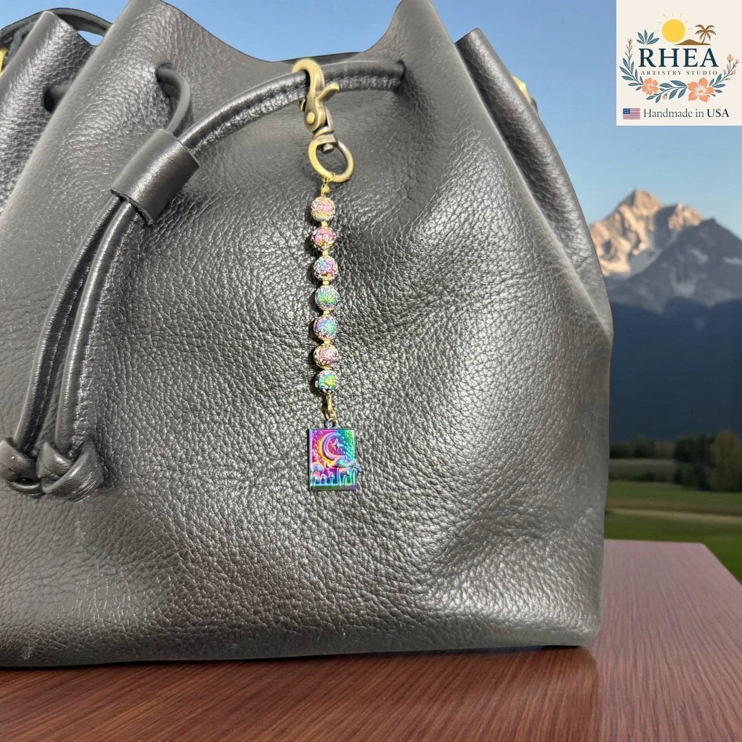 Rhea Artistry Studio rainbow charm with moon and cityscape pendant, beaded with iridescent glass and gemstones, attached to a black leather bag.