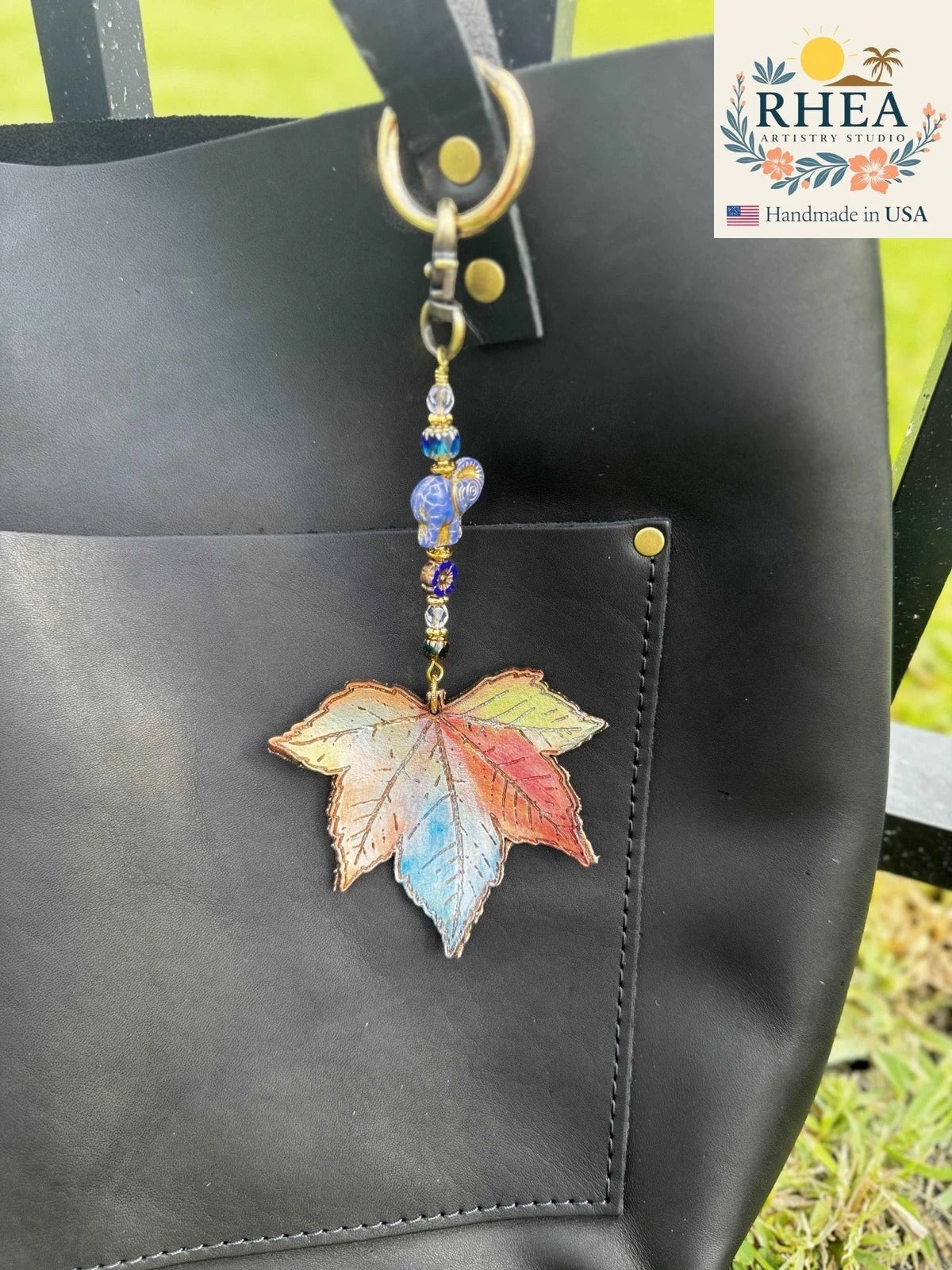 Rhea Artistry Studio rainbow elephant charm with a hand-painted leather leaf, Czech glass beads, and brass hardware, attached to a black leather tote bag.