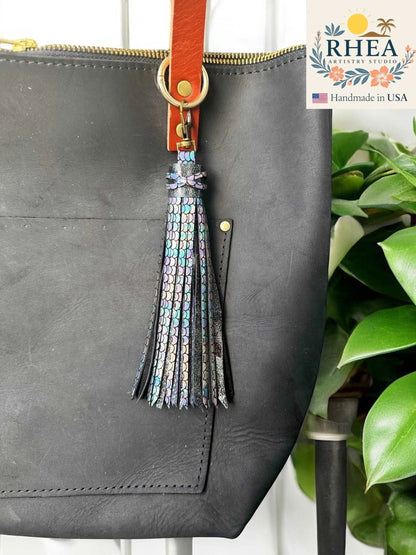 Rhea Artistry Studio 7-inch rainbow chameleon leather tassel keychain with lobster clasp attached to a black leather tote bag.